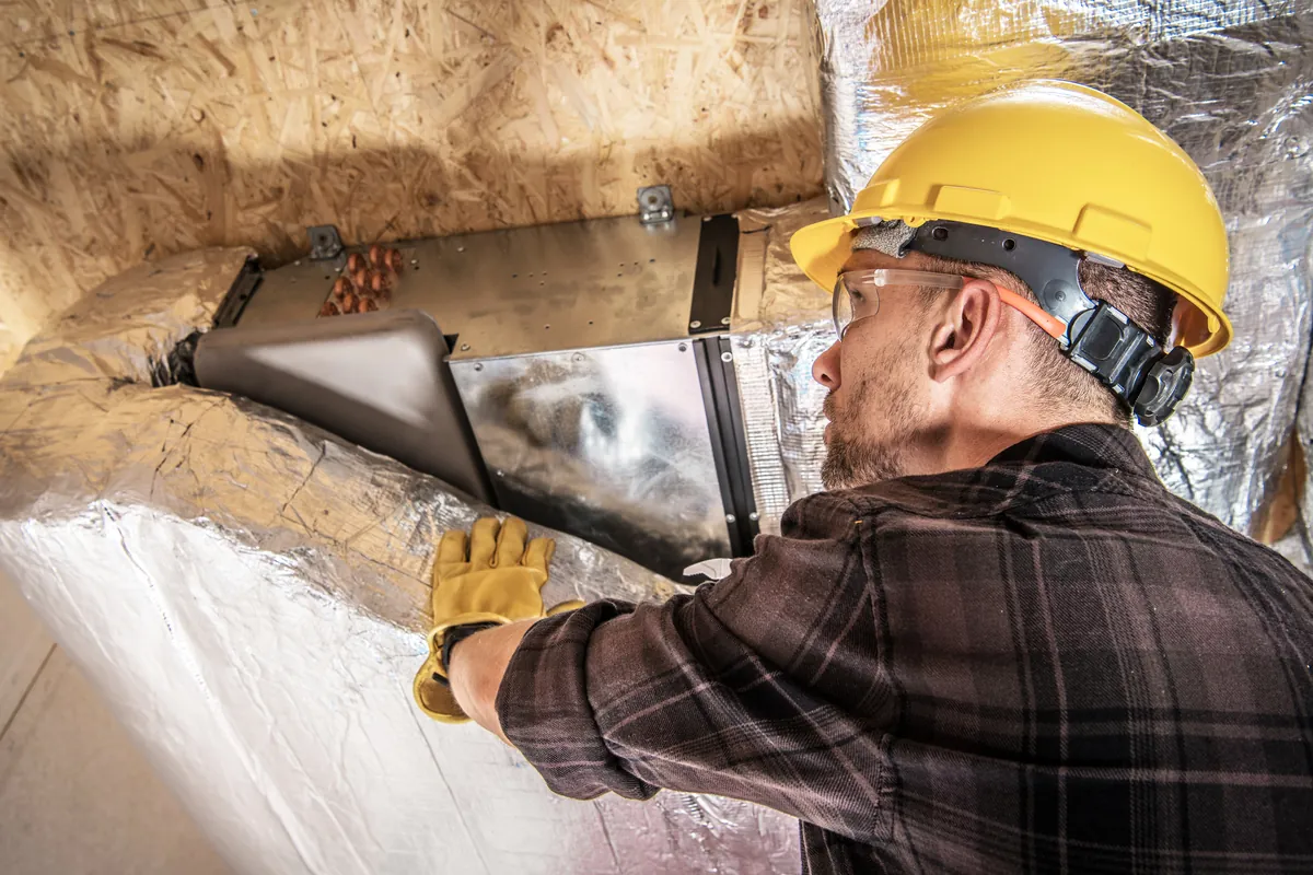 A professional HVAC technician in protective gear repairing foil-wrapped air ductwork in an unfinished Los Angeles building.