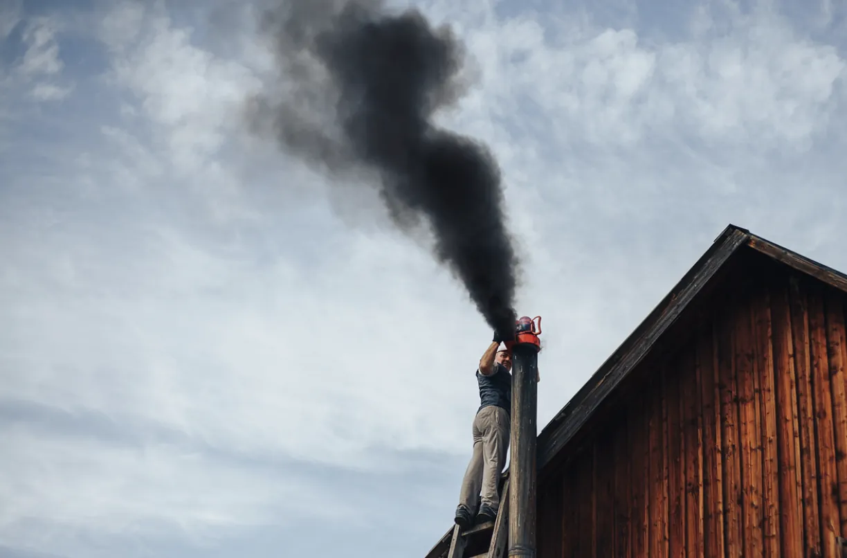 Professional chimney sweep in the San Fernando Valley climbing a ladder to inspect a residential flue releasing heavy black smoke, indicating a severe creosote buildup or blockage.