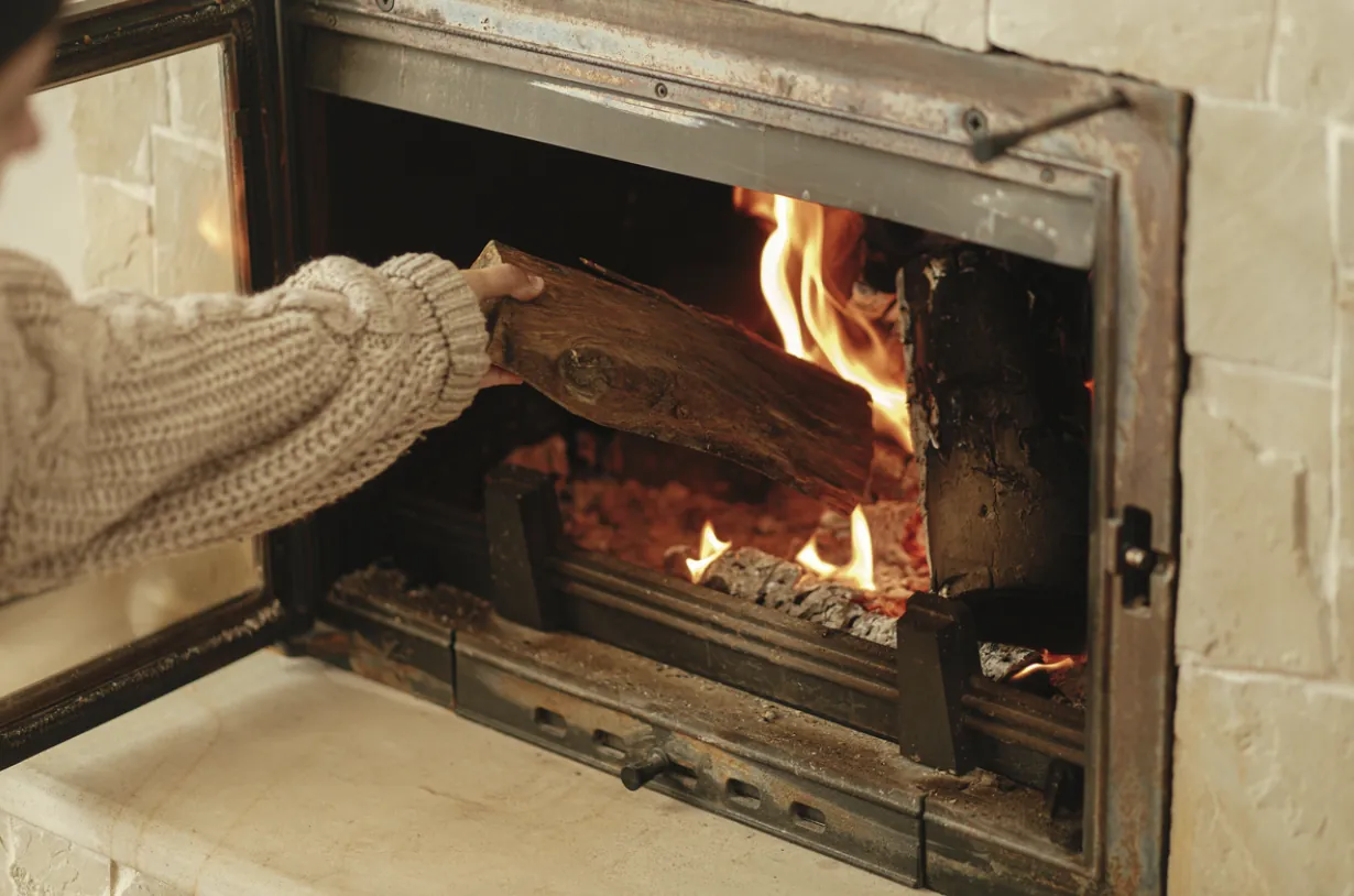 Hand placing a log into a clean, beige stone fireplace in a Los Angeles home, highlighting the importance of regular soot removal and hearth maintenance for safe wood-burning.
