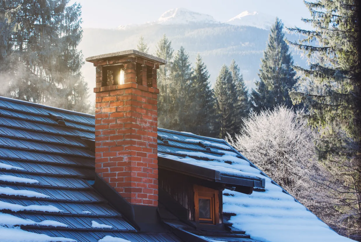 Residential brick chimney on a snowy rooftop with frost-covered trees and misty mountains in the background, illustrating the importance of annual winter chimney inspections in Los Angeles.
