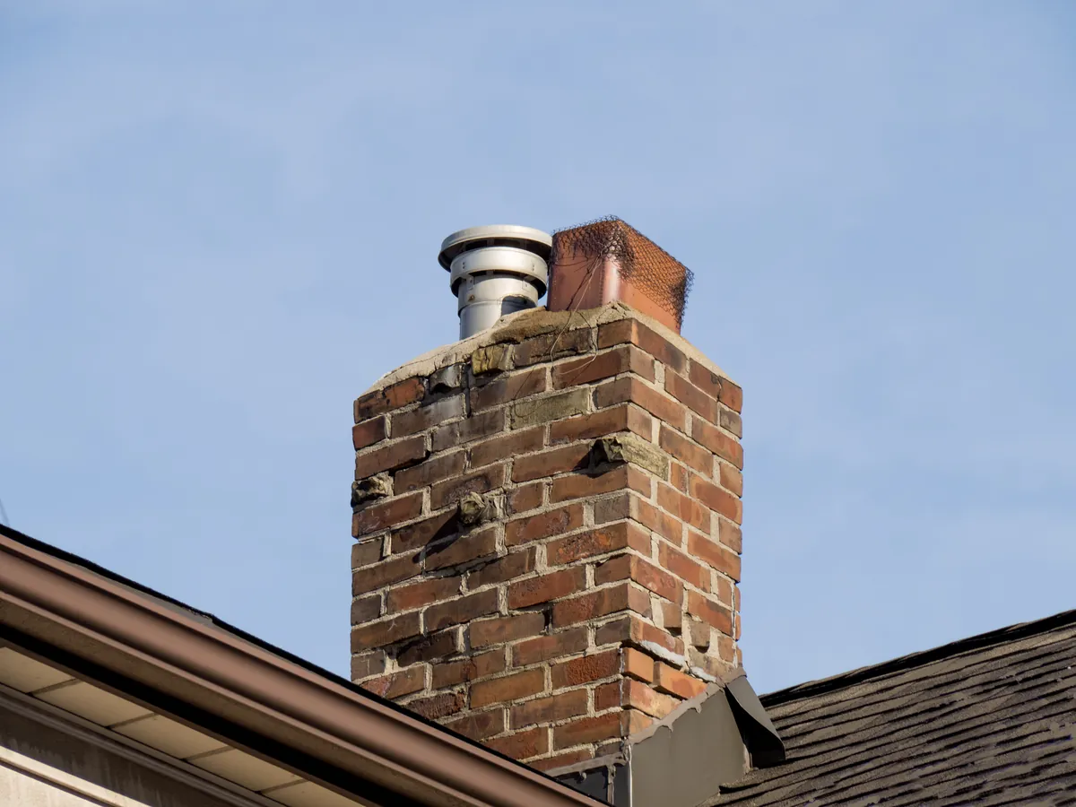 A weathered red brick chimney with a metal cap on a shingled roof, requiring professional leak inspection and masonry repair in Los Angeles.